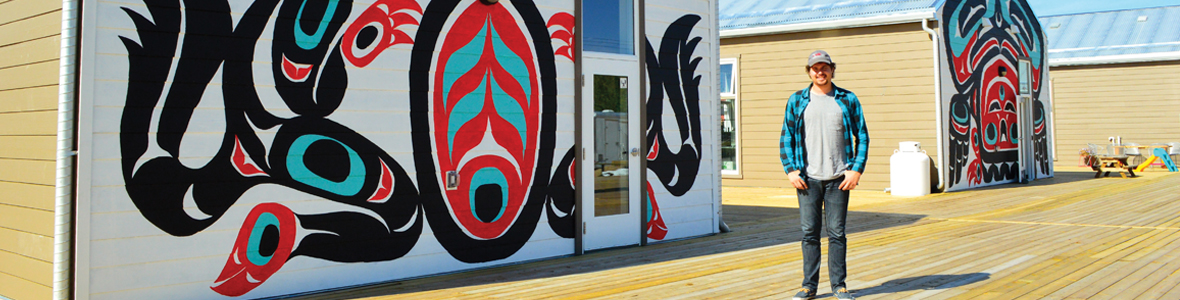Male CO-OP student posing for a picture near a mural with indigenous art in Whitehorse, Yukon